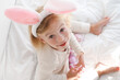 Happy caucasian girl two years old wearing rabbit bunny ears playing with eggs sitting on bed looking at camera. Easter holiday celebrating.High angle view shot.