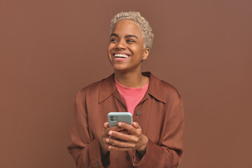 Wall Mural - Young joyful casual African American woman with phone in hand looks up and laughs feeling improvement in mood after reading positive news or SMS messages stands on brown background