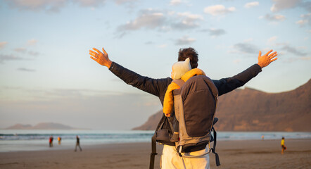 Wall Mural - Young father rising hands to the sky while enjoying pure nature carrying his infant baby boy son in backpack on windy sandy beach of Famara, Lanzarote island, Spain at sunset. Family travel concept