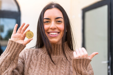 Poster - Young woman holding a Bitcoin at outdoors pointing to the side to present a product