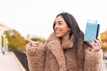 Poster - Young woman holding a passport at outdoors pointing to the side to present a product