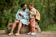 © Seventyfour - Full length portrait of black mother and daughter sitting on rock while hiking together in nature, copy space