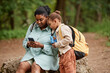 © Seventyfour - Portrait of black mother and daughter sitting on rock while hiking together and using mobile phone
