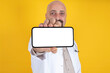 © Designerant - Male doctor showing big horizontal smartphone with white blank screen for mock up. Standing over yellow background. Hospital professional medical mobile application or website or advertising concept.
