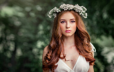 Wall Mural - Beautiful red haired girl in white vintage dress and wreath of flowers standing under the tree and looking at camera. Fairytale story. Warm art work.