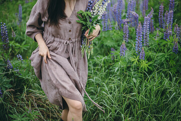 Wall Mural - Close up of woman in rustic dress holding lupine bouquet in meadow. Cottagecore aesthetics. Young female in linen dress walking with wildflowers in atmospheric summer countryside, rural slow life