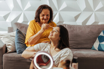 Smiling grandmother applying makeup on her granddaughter. Candid moment of affection between grandmother and adult granddaughter.