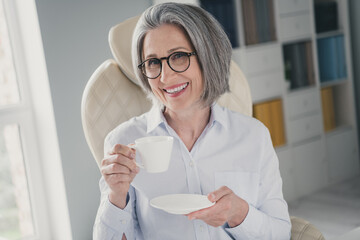 Sticker - Photo of cheerful successful aged lady sitting leather chair arm hold enjoy fresh coffee mug modern office inside