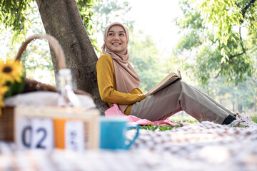 Photo of muslim girl student wearing headscarf sitting on blanket in green park and reading book under big tree. Picnic concept