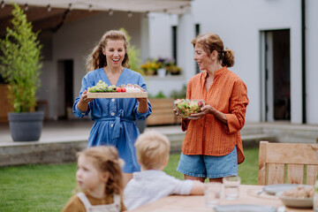 Wall Mural - Happy daughter and mother bringing salad and burgers at multi generation garden party in summer.