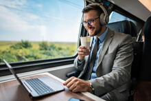 Handsome Businessman Is Having A Good Time While Traveling By High-speed Train. He Is Using Laptop Computer And Wireless Headphones For Online Communication, Gaming And Entertainment.
