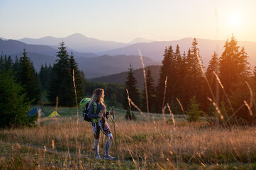 Woman traveller hiking outdoors at sunset. Sporty, slim woman traveling in mountains. Female tourist carrying backpack, using trekking sticks, admiring landscape. Concept of harmony with nature.