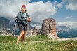 Smiling female trekker with backpack and trekking poles on green mountain hill enjoying picturesque Dolomite Alps. Cinque Torri formation on the background. Active people and mountains concept.