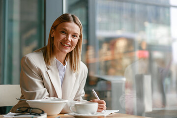 Wall Mural - Smiling businesswoman having lunch and making notes while working in cafe