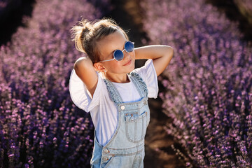 Wall Mural - adorable child girl in lavender field on sunset. smiling kid in sunglasses, jeans jumpsuit is having fun on nature on summer day. Family day, vacation, holiday.International Children's Day,