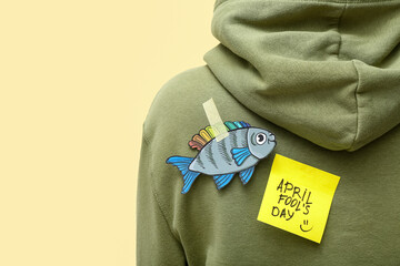 Wall Mural - Sticky paper with text APRIL FOOL'S DAY and fish on woman's back against color background, closeup