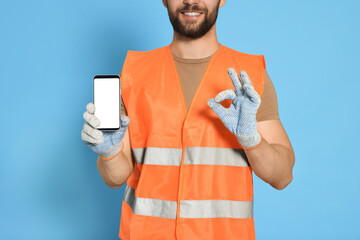 Canvas Print - Man in reflective uniform showing smartphone and OK gesture on light blue background, closeup