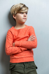 A serious boy of 9-10 years old stands near a gray wall. The guy in the orange sweater crossed his arms over his chest. Vertical.