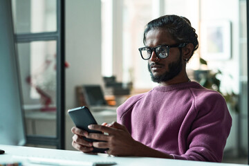 Young programmer in eyeglasses using mobile phone in his online work at table