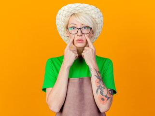 Wall Mural - young gardener woman with short hair in apron and hat pointing with index figners at her eyes looking at camera confused standing over orange background