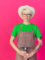 Wall Mural - young gardener woman with short hair in apron and hat holding rake lookign at camera smiling confident standing over pink background