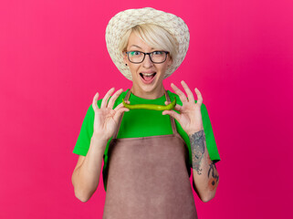 Wall Mural - young gardener woman with short hair in apron and hat holding green chili pepper looking at camera smiling cheerfully standing over pink background