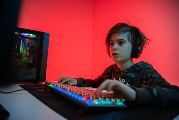 Wall Mural - Image of immersed teenage gamer boy playing video games on computer in dark room wearing headphones and using backlit colorful keyboard