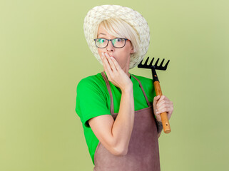 Wall Mural - young gardener woman with short hair in apron and hat holding mini rake looking at camera being amazed and surprised standing over light background