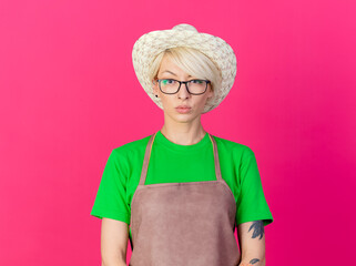 Wall Mural - young gardener woman with short hair in apron and hat looking at camera with serious face standing over pink background