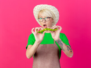 Wall Mural - young gardener woman with short hair in apron and hat holding green chili pepper breaking it looking surprised standing over pink background