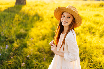 Wall Mural - Portrait of lovely redhead young woman in straw hat and white dress standing posing on beautiful meadow of green grass looking at camera, on background of warm sunlight at summer day.