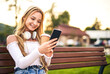 © PEOPLEVISUALS - Young happy teenager girl sitting on a bench and chatting on a smart phone while holding coffee to go paper mug in her hand - Mobile technology concept