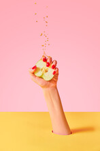 Closeup Female Hand With Red Painted Nails Squeezing Half Of Apple Fruit Over Pink Yellow Background. Contemporary Art. Drops Of Juice Fly Up