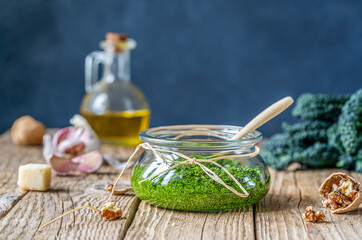 Wall Mural - Homemade kale pesto served in a glass jar on wooden surface with dark background and basic ingredients - kale, walnuts, parmesan, garlic and olive oil