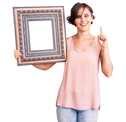 Canvas Print - Beautiful young woman with short hair holding empty frame surprised with an idea or question pointing finger with happy face, number one