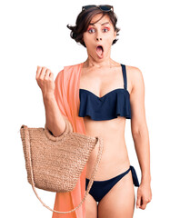 Poster - Beautiful young woman with short hair wearing bikini and hat holding summer wicker handbag scared and amazed with open mouth for surprise, disbelief face