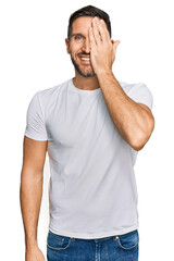 Wall Mural - Handsome man with beard wearing casual white t shirt covering one eye with hand, confident smile on face and surprise emotion.