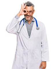 Sticker - Middle age grey-haired man wearing doctor uniform and stethoscope smiling happy doing ok sign with hand on eye looking through fingers