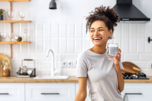 Portrait of a beautiful African American young woman drinking water standing in the kitchen at home looking away and smiling, wellness healthy food concept