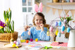 © ArtSys - Portrait of cute pretty little girl in blue dress and funny rabbit ears making craft, diy eggs for easter holiday home decoration. Spring colours, bright flowers. Kid's domestic life, hobby, enjoyment