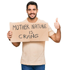 Wall Mural - Young hispanic man holding mother nature is crying protest cardboard banner smiling happy and positive, thumb up doing excellent and approval sign