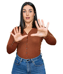 Wall Mural - Young hispanic woman wearing casual clothes afraid and terrified with fear expression stop gesture with hands, shouting in shock. panic concept.