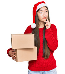 Wall Mural - Young chinese woman wearing christmas hat holding delivery package serious face thinking about question with hand on chin, thoughtful about confusing idea