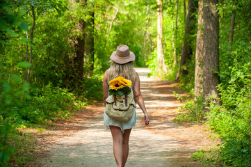 A blonde caucasian woman walks down a trail in the forest with sunflowers sticking out of her backpack on a sunny Summer's Day 