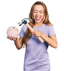 Poster - Young caucasian woman holding piggy bank and hammer sticking tongue out happy with funny expression.