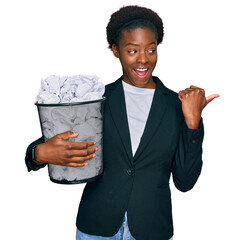 Sticker - Young african american girl holding paper bin full of crumpled papers pointing thumb up to the side smiling happy with open mouth