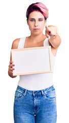 Sticker - Young beautiful woman with pink hair holding empty white chalkboard pointing with finger to the camera and to you, confident gesture looking serious