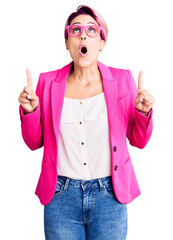 Canvas Print - Young beautiful woman with pink hair wearing business jacket and glasses amazed and surprised looking up and pointing with fingers and raised arms.