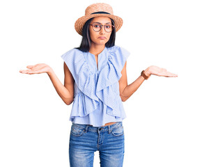 Sticker - Young beautiful latin girl wearing summer hat and glasses clueless and confused expression with arms and hands raised. doubt concept.