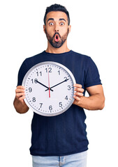 Wall Mural - Young hispanic man holding big clock scared and amazed with open mouth for surprise, disbelief face